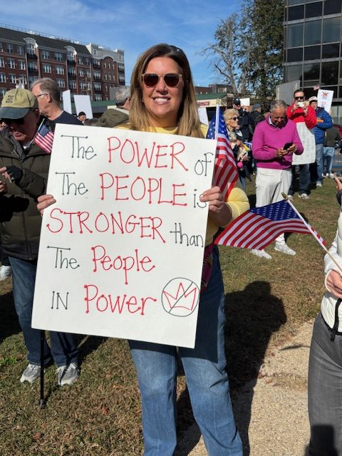 A smiling woman holds a handmade sign saying, "The POWER of the PEOPLE is STRONGER than the People in Power." A hand-drawn "No Kings" symbol is on the bottome corner. 