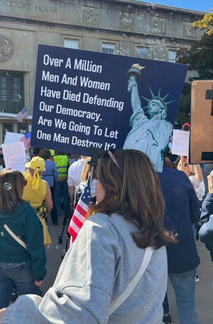 A woman holds up a sign with an image of the Statue of Liberty. The text reads, "Over a million men and women have died defending our democracy. Are we going to let one man destroy it? 