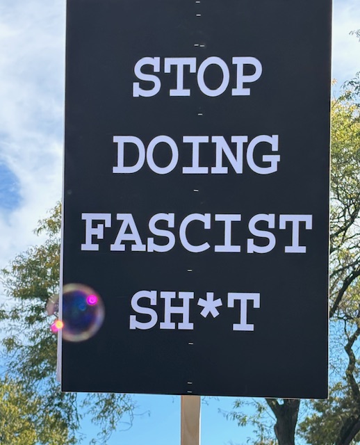 A sign on a black background says, "STOP DOING FASCIST SH*T" 
