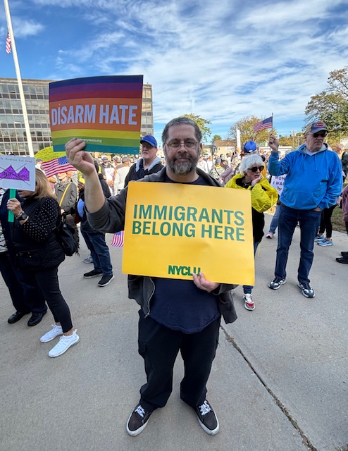 A man holds two signs from the NYCLU. One yellow one says, "IMMIGRANTS BELONG HERE." The other has rainbow colors and says, "DISARM HATE."