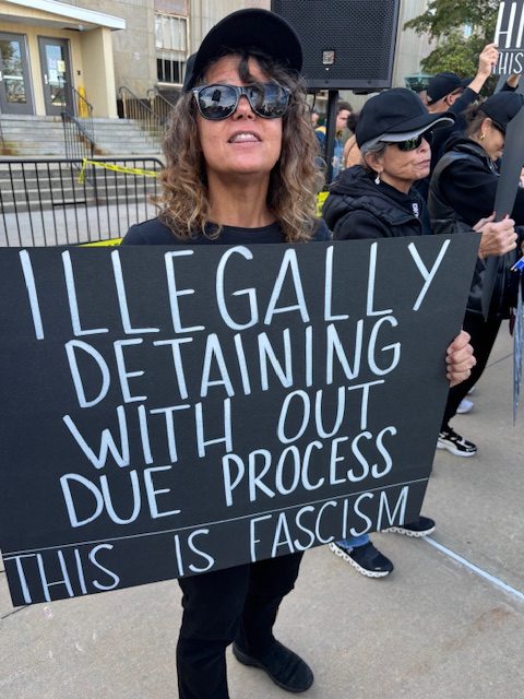 A woman holds a handmade sign that says, "ILLEGALLY DETAINING WITH OUT DUE PROCESS -- THIS IS FASCISM"
