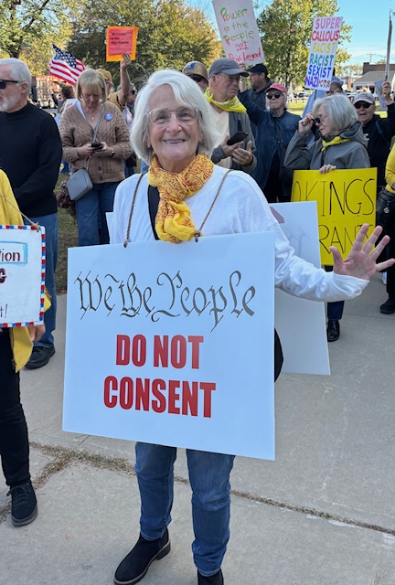 An older woman wearing a yellow scarf holds a sign that says, "We the People DO NOT CONSENT"