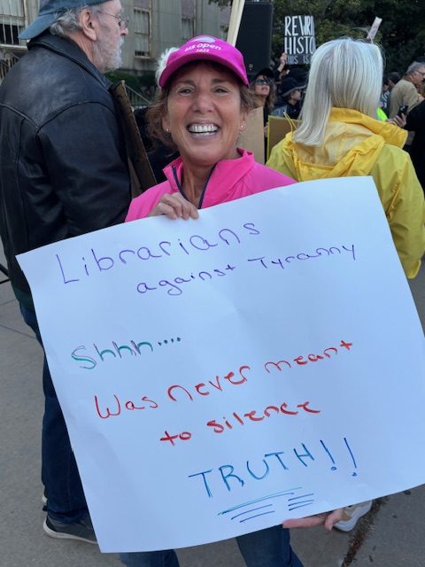 A librarian holds up a handmade sign that says, "Librarians against Tyranny. Shhh... Was never meant to silence Truth!!"